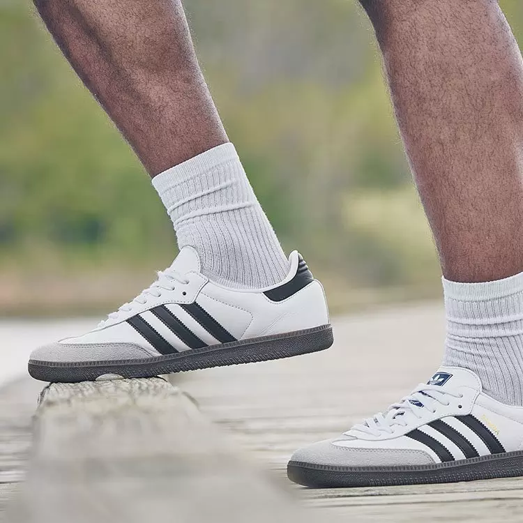 White adidas Samba sneakers with black three-stripes and grey suede toe caps worn with white ribbed crew socks while walking on a wooden boardwalk.