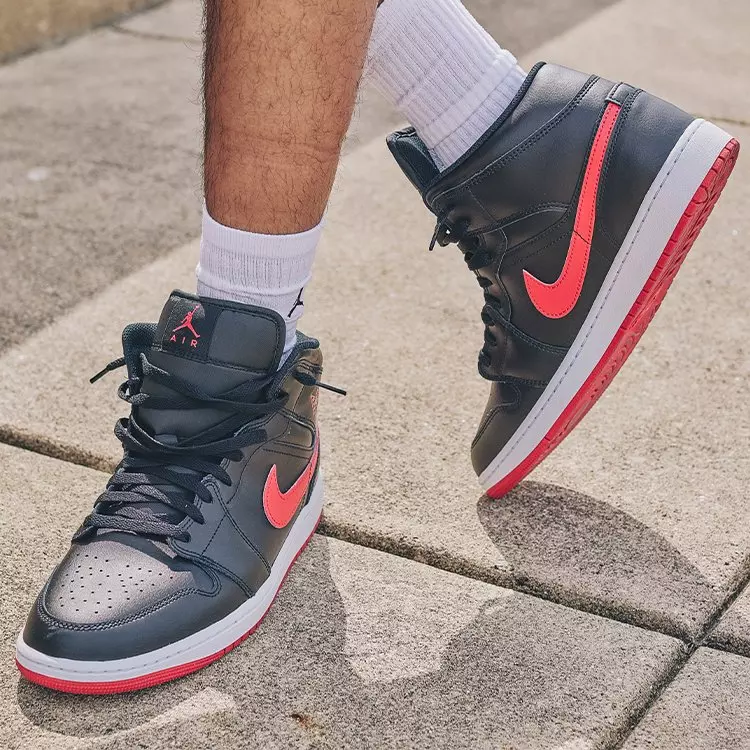 Black Air Jordan 1 Mid sneakers with bright red swooshes and matching red outsoles, worn with white Jordan crew socks while walking on a concrete sidewalk.