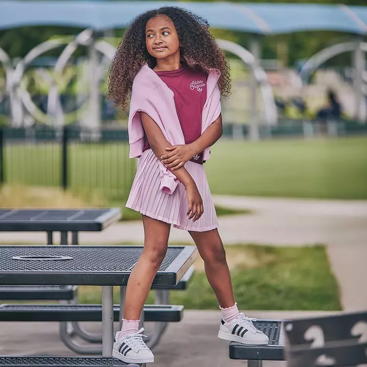 A young girl poses outdoors on a park table wearing a maroon adidas t-shirt, light pink pleated skirt, draped hoodie, and white Superstar sneakers.